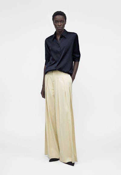 Μαύρη γυναίκα που στέκεται με τα χέρια στις τσέπες φορώντας μπλε σατέν πουκάμισο και κρεμ πλισέ παντελόνι με φαρδύ πόδι σε λευκό φόντο.