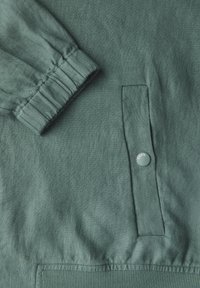 Detalle de chaqueta de lino verde con puño fruncido y bolsillo con solapa y botón a presión marcado con "CECIL". Tejido texturizado con un acabado suave.