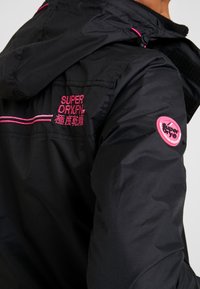 Schwarze, leichte Jacke mit Kapuze. Mit rosa gesticktem Branding und Akzenten. Strukturierter Stoff mit glatter Oberfläche.