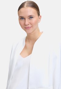 Blazer blanc au toucher lisse et au design minimaliste, porté sur un haut blanc. L'arrière-plan est simple et clair.