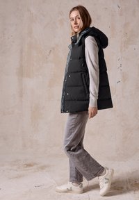 Schwarze Steppweste mit Kapuze, mit Druckknöpfen und gestepptem Muster, kombiniert mit grauen Jeans und weißen Sneakers mit grünen Akzenten.