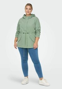 Šviesiai žalia su gobtuvu striukė su susiaurinta juosme, dviem priekinėmis kišenėmis ir reguliuojamais raišteliais. Sujungta su mėlynais džinsais ir baltais sportbatiais.