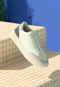 Baskets blanches et grises avec des lacets flottant au-dessus de marches crème et rouges contre un mur bleu.