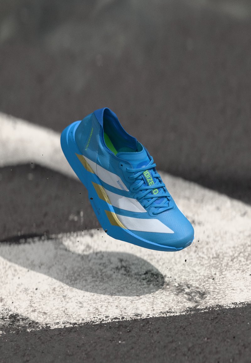 Ελαφρύ παπούτσι τρεξίματος σε έντονο μπλε χρώμα με λευκές και χρυσές λεπτομέρειες. Διαθέτει αναπνεύσιμη σχεδίαση και υφασμάτινη σόλα για πρόσφυση, αιωρούμενο πάνω από έναν στίβο.