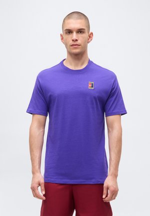 Mand med kort hår iført en lilla t-shirt med rund hals og et lille flerfarvet kvadratisk logo på brystet samt røde shorts, vendt fremad.