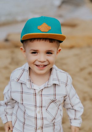 Cappellino turchese e giallo con un logo, camicia a quadri bianchi con accenti e bottoni marroni. Bambino con un'espressione allegra su uno sfondo sabbioso.