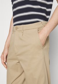 Pantalon en coton beige à texture lisse, équipé d'une poche avant, d'une fermeture par bouton et d'une coupe ajustée. Porté avec un haut rayé bleu marine et blanc.