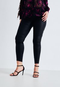 Leggings en simili cuir noir avec une coupe ajustée, associés à des sandales à talons hauts noires à lanières. Le blouse présente un motif floral en rose.