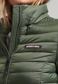 Närbild av en person som bär en grön Superdry quiltad jacka med hög krage och logotypmärke på bröstet.