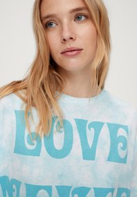 Lichtblauw tie-dye t-shirt met grote, vetgedrukte turquoise tekst die "LOVE" zegt, met een ontspannen, oversized pasvorm en zachte katoen.