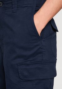 Main dans la poche d'un pantalon cargo bleu marine avec poche à rabat latérale et détails de couture visibles.