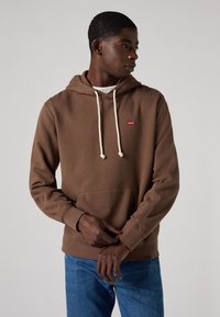 Felpa marrone in misto cotone, con cappuccio regolabile, tasca frontale e un piccolo logo rosso sul petto, abbinata a jeans blu.
