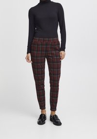 Rode en groene geruite broek met plooien, gemaakt van een zachte stof, gecombineerd met zwarte loafers en een aangesloten zwarte coltrui.