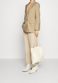 Blazer beige con botones dorados, pantalones beige claros, bolsa de mano de piel blanca, botas tobilleras blancas texturizadas con punteras puntiagudas y tacones pequeños.