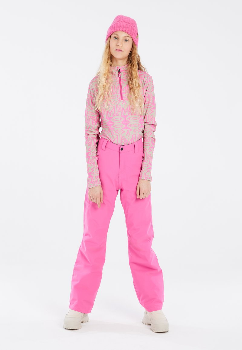 Pantalons de ski roses et un haut à manches longues assorti avec un motif géométrique. Le mannequin porte un bonnet en tricot rose et des bottes beiges.