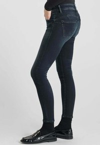 Jeans skinny de mezclilla oscura con un diseño ajustado, cintura alta y costuras mínimas. Llevados con mocasines negros y pantys, mostrando una silueta elegante.
