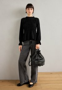 Haut noir à manches longues froncé avec un col à volants, associé à un jean gris à jambes larges et un sac seau noir texturé. Ballerines noires.