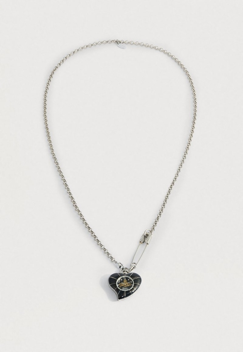 Sudraba ķēdes kaklarota ar melnu sirds formas piekariņu, kas ir rotāts ar zelta detaļām, un spīdīgu drošības adatu piekariņa tuvumā.