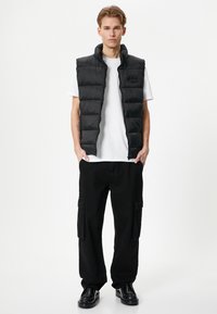 Schwarze Pufferweste mit hohem Kragen und horizontalen Steppnähten, getragen über einem weißen T-Shirt; kombiniert mit schwarzen Cargo-Hosen und Schuhen.