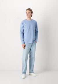 Felpa azzurro chiaro a maniche lunghe con texture a coste, abbinata a jeans a gamba dritta in denim chiaro e sneakers bianche, in posa davanti a uno sfondo bianco.