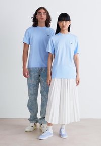 Himmelblaue Baumwoll-T-Shirts im lockeren Schnitt, kombiniert mit einem weißen, gefalteten Rock und camo-musternden Hosen. Die Accessoires umfassen blaue Turnschuhe.