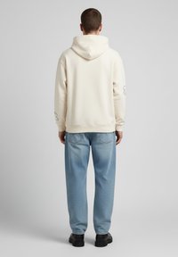 Felpa crema con cucitura sul retro e un sottile dettaglio grafico sul braccio sinistro, abbinata a jeans azzurri e scarpe nere.