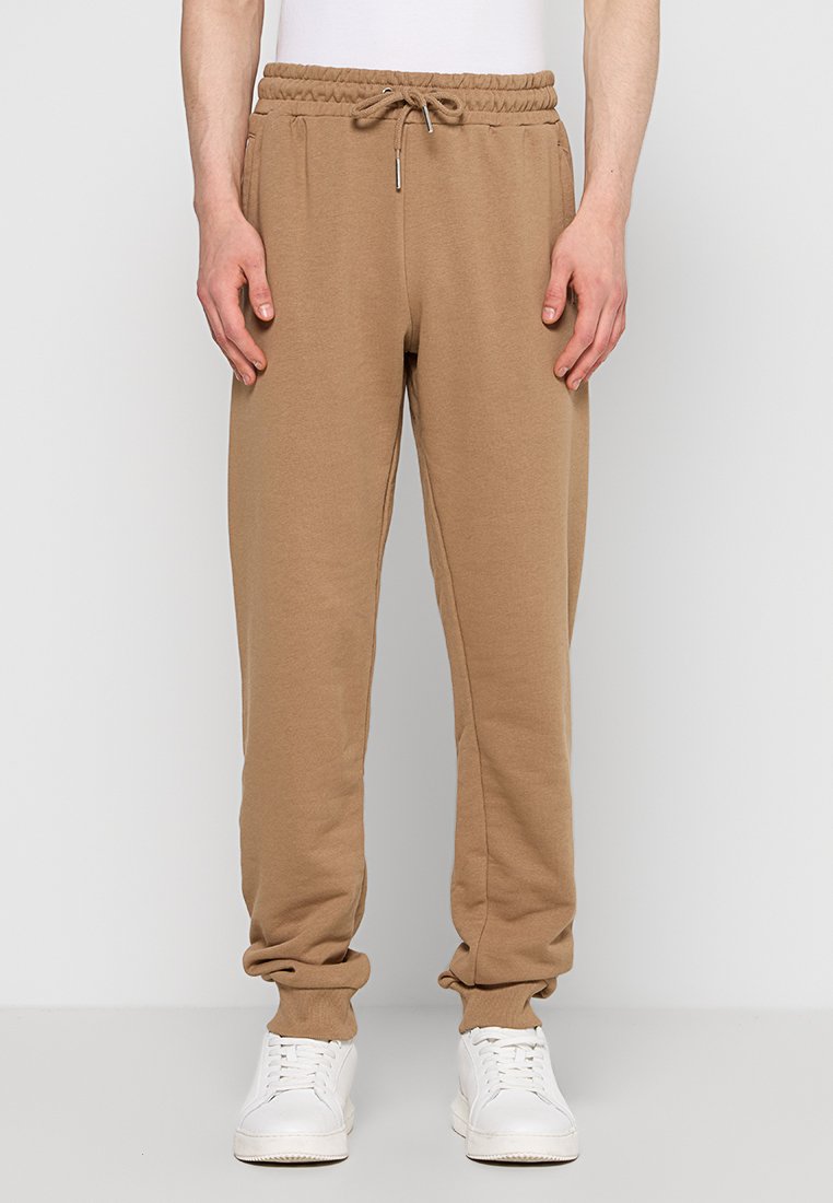 Pantaloni sportivi in cotone marrone con vita elastica, dettaglio con cordoncino e caviglie a costina. Indossati con sneakers bianche.