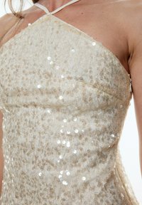 Halterkleid aus hellbeige Pailletten mit transparenter Textur, ausgestattet mit kleinen runden Pailletten und einer figurbetonten Silhouette.