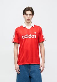 Νέος άνδρας που φοράει κόκκινο πόλο της Adidas με λευκές ρίγες στα μανίκια και μπλε τζιν, στέκεται μπροστά σε απλό λευκό φόντο.