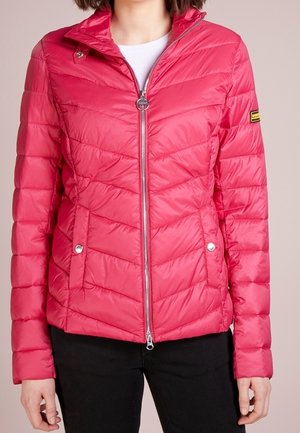 Frau trägt eine leuchtend rote, gesteppte Jacke mit Frontreißverschluss, Druckknopftaschen und schwarze Hose vor neutralem Hintergrund.