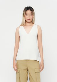 White, sleeveless top with a deep V-neck, textured fabric, straight hem, and minimal seams, paired with khaki pants.