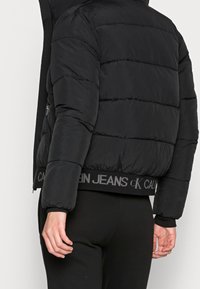Giacca a vento nera con cuciture orizzontali e bordo a costine. Presenta una fascia con logo in vita e chiusura con zip sul lato. Tessuto testurizzato.