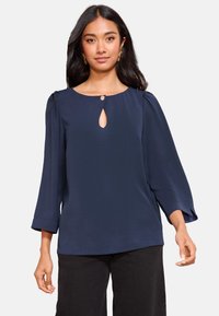 Marineblauwe blouse met driekwart mouwen, een ronde halslijn en een sleutelgatuitgesneden detail met een kleine knoop. Gestructureerde stof met een ontspannen pasvorm.