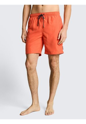 Mann, der leuchtend orange Badeshorts mit schwarzer Kordel trägt, barfuß vor einem schlichten hellen Hintergrund steht.