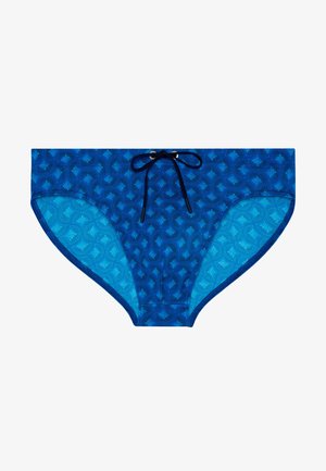 Modré pánské plavky ve stylu slipů s tmavě modrým geometrickým vzorem a černým stahovacím páskem v pase, položeny na bílé ploše.