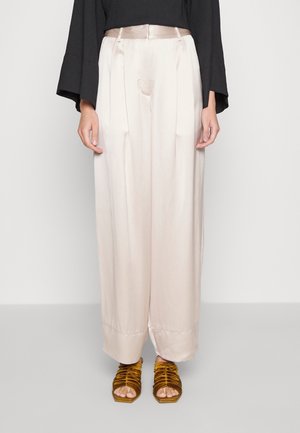 Weit geschnittene, blassrosa Satin-Hosen kombiniert mit senffarbenen, gerafften Sandalen mit offenen Zehen und einem schwarzen Langarmshirt, von der Taille abwärts gezeigt.