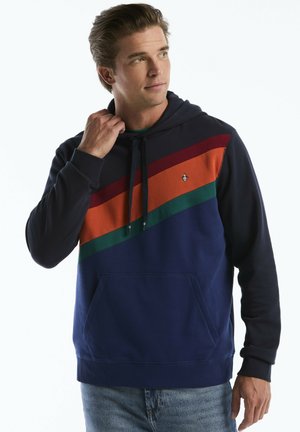 Man wearing navy hoodie with diagonal green, orange, and burgundy stripes, holding the hood string with one hand and looking sideways.