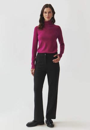 Maglione fucsia con collo a imbuto abbinato a pantaloni neri a gamba larga. I pantaloni presentano un bottone frontale e l'outfit è completato da scarpe nere.