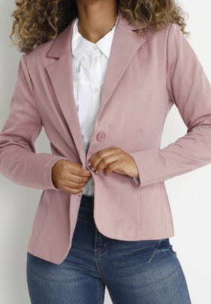 Frau mit lockigem Haar, die einen pinken Blazer über einem weißen Hemd mit Knöpfen und dunkelblauer Jeans schließt.