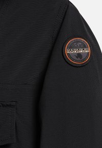 Rękaw czarnej kurtki z okrągłą naszywką marki Napapijri z pomarańczową obwódką, ikoną globu i napisem na górnej części ramienia.