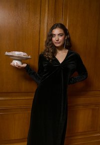 Svart sammetsklänning med V-ringning och långa ärmar, med en vridningsdetalj vid bysten. Modellen håller en vit kakfat med dessert.