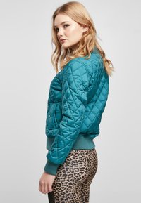 Teal quiltad bomberjacka med ribbad nederkant och muddar. Har en framficka och en slät, glänsande textur. Bärs med leopardmönstrade leggings.