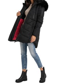 Svart vadderad vinterjacka med pälsfodrad huva, öppnad för att avslöja en kontrasterande röd inredning, tillsammans med blå jeans och svarta ankelstövlar.