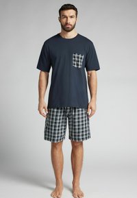 Camiseta de algodón azul marino de manga corta con un bolsillo en el pecho de cuadros, combinada con pantalones cortos de cuadros en azul marino, negro y blanco. Descalzo.