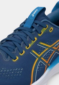Chaussure de sport bleue avec tige en maille, accents orange et jaune, lacets plats et semelle amortie. Présente une languette bleue sur le dessus.