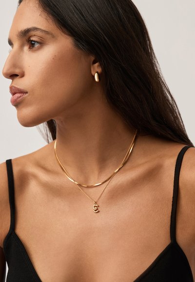 Γυναίκα με μακριά σκούρα μαλλιά, φορώντας μαύρο τοπ, χρυσό κρίκο σκουλαρίκι, στρωμένα χρυσά κολιέ, κοιτάζοντας προς τα αριστερά της σε ουδέτερο φόντο.