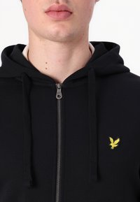 Hanorac negru cu fermoar confecționat dintr-un material moale, având un fermoar frontal și un logo brodat galben pe partea stângă a pieptului.