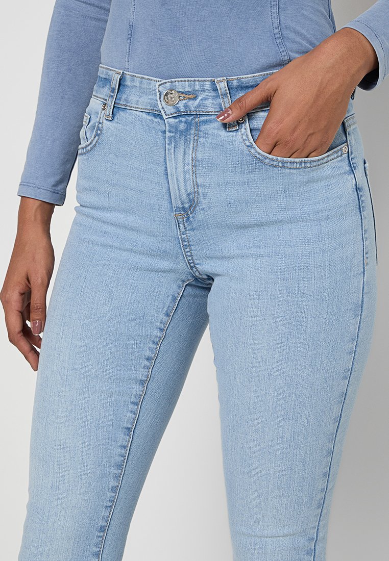 Person in einer hellblauen, hoch taillierten Jeans mit der Hand in der vorderen Tasche, kombiniert mit einem hellblauen Langarmoberteil.