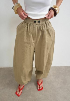 Woman wearing beige wide-leg pants with buttons, a white sleeveless top, red thong sandals, and multiple chunky bracelets on both wrists.
