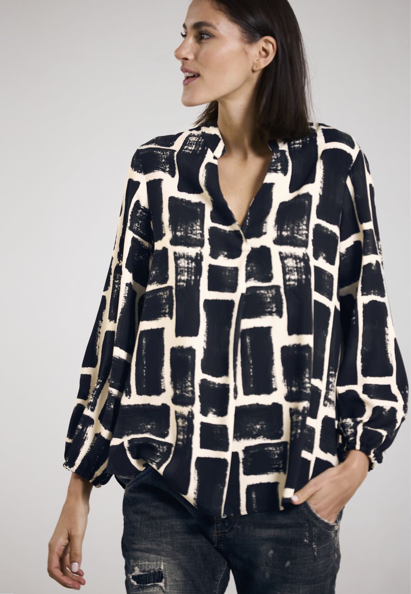 Schwarze und cremefarbene gemusterte Bluse mit lockerer Passform, langen Ärmeln und V-Ausschnitt, versehen mit abstrakten Pinselstrich-Quadratdesigns.